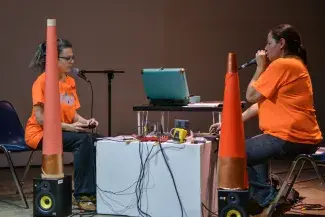 Two women, wearing blue jeans and orange t-shirts, sit across from each other, between them a white plinth is covered with sound equipment. Each is speaking into a microphone as they mix their voices. Near their feet there are two speakers with large orange paper traffic-cone-esque objects sitting on top of them which partially obscure the artists from view. 
