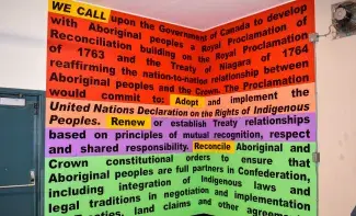The words WE CALL, written in black text and highlighted in yellow, looms at the top of a hallway corner wall which is fully painted in text. The text is highlighted in green, salmon pink, purple and green and each segment is started with a verb: Adopt, Renew, Reconcile. From top to bottom the text reads in part: WE CALL upon the Government of Canada to develop with Aboriginal peoples a Royal Proclamation of Reconciliation building on the Royal Proclamation of 1763 and the Treaty of Niagara of 1764
