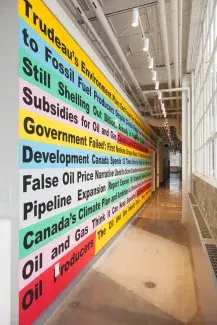 Black bold text colourfully highlighted in yellow, light blue, teal, grey and red runs down the length of a long cement hallway. The painted text brightened by the light streaming in the adjacent windows can be partially read: Trudeau’s Environment Plan: End Fossil…to Fossil Fuel Producers Despite Climate Pledge…Still Shelling Out Billions Annually…Subsidies for Oil and Gas, Canada leads G7…Government Failed: First Nations Groups…Development Canada Spends 12 times…False Oil Price Narrative Used to Scare…