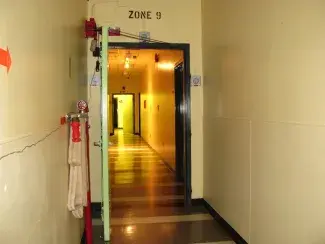 A flash photograph exposes the khaki painted concrete walls of a long bunker hallway equipped with a fire hose secured to the left wall. A stencilled sign reads Zone 9 above an open green metal door through which the hallway extends cast in yellow fluorescent light which evokes an auditory hummm.