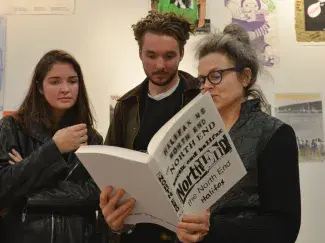 Cathy, a woman with grey hair tied in a bun and dark rimmed glasses, reads aloud from The North End Halifax, a large white paper covered book. Two viewers look over her shoulder at the pages. Behind them posters cover the white walls which reflect the posters found in the publication. 