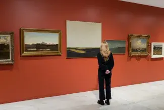 A viewer stands in front of a large landscape painting with a vast grey sky and dark green rolling grasses. The surrounding paintings are shifted up and down on the wall inorder to align the horizon lines of each neighbouring painting. 
