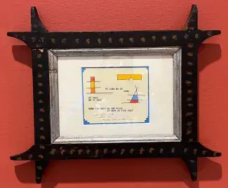 A small note paper framed with blue border holds orange and yellow shapes and the words quote: su casa es mi casa, mi casa es su casa, when you shit on the floor it gets on your feet, are written using a typewriter. The whole note is framed with black wood that has swooping pointed corners which look hand carved.