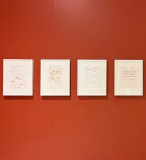 Four framed documents ranging from note paper drawings to typed letters hang in a row on a vermillion wall. 