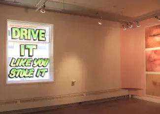 Sunlight streams into a gallery space through a large window and the words DRIVE IT LIKE YOU STOLE IT are painted in black and green graffiti style font across the glass. On the right hand wall three prints of mouths are partially visible, each one cropped and enlarged showing the frozen expression in detail; lips mid-way through a word, just about to speak or pursed shut in a word swallowing frown.