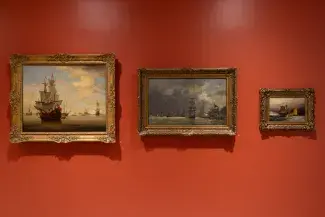 Three ocean landscape paintings are hung against a vermillion wall. Each has a golden filigree frame which decreases in size from left to right. The horizon lines of each different sea and sky align although the composition of ships and stormy or calm waters ranges. 