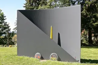  A large grey metal sculpture protrudes from a field of grass. Two circular discs with red and yellow designs lean against a right-angled triangular sheet of metal which connects and wraps into a curved rectangular slab. The small end of a long yellow megaphone pokes out from between the metal shapes.