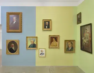 Nine portraits hang against dusty blue, wheaty yellow,  and pale green wall panels. The grey blues of ageing men’s hair tone with silk garments and yellow and green undertones surface between various skin shades. 