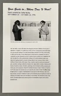 A thin xerox printed pamphlet titled Your guide to…Where Does It Hurt? curated by Cathy Busby September 20 - October 18 1996, rests on a grey background. There is a black and white picture of two figures reading books in front of Cathy Busby’s Self-Help Library below the title with a long description that begins: Pain, like death, is hard to talk about even though we all know it.