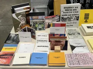 A table loaded with an array of colourful publications by artist Cathy Busby. Some books lie flat, like Sorry and Branded, while other pamphlets stand up partially opened, like Beijing-Vancouver.  