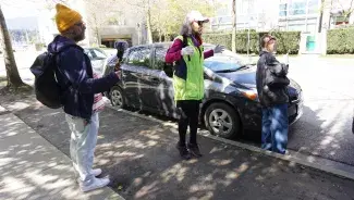 Three people are standing on the sidewalk near an urban street. Cathy, wearing a bright green vest and pink baseball cap over her grey hair, stands in between a man with a yellow toque and grey beard holding an audio recorder and a young person with a buzz cut taking a picture of the model of the car parked on the street next to them. Cathy is standing on her tip toes with her hands in the air like a conductor, her lips are pursed mid sentence, mid song. 