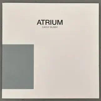 A plain grey-blue rectangle sits on the left parameter of a small square cardstock folder, the colour block wraps around the side of the folder encouraging the reader to turn it over or open it up. The rest of the cover is white. The works title, Atrium, is written in black sans serif font on the upper centre, and the artist's name, Cathy Busby, is written in a smaller font just below. 