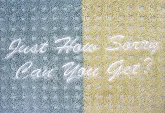 A two toned towel, half light blue and half light yellow, has the question Just How Sorry Can You Get? stitched onto it in cursive lettering in white thread.