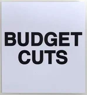 A white square of cardstock is mostly filled with the words BUDGET CUTS  written in bold black sans serif font. 