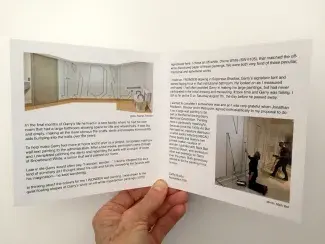 A soft hand holds open a cardstock folder. A panorama image of a washroom mid way through pre-paint tapping tops paragraphs of text, partially reading: In the final months of Garry’s life he lived in a care facility where he had his own room that had a large bathroom allowing space for lists and wheelchairs. It was big and empty, making all the more obvious the scuffs, dents, and scrapes from mobility aids bumping into the walls over the years…Cathy Busby, November 2021. 