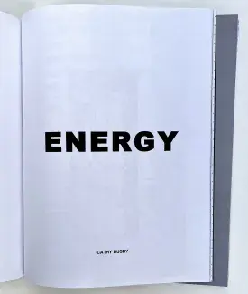 An open book of thin letter paper. ENERGY is written in bold black letters across the right page, Cathy Busby is written in smaller font at the bottom of the same page. Inky shadows seep through from an indiscernible black and white image on the other side. 
