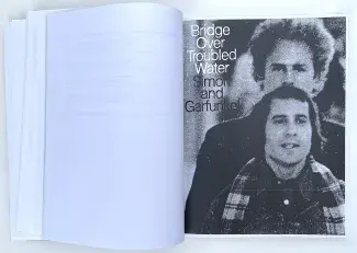 An open book, the left page is blank legal paper, the right page is a xeroxed black and white photo of Simon and Garfunkel's Bridge Over Troubled Water album cover: a grainy image of Simon, with his brown hair in a bob, standing in front of Garfunkel, who’s blond curls halo his head, with the album title and their names in contrasting font beside their faces. 