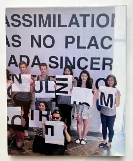 An image of students standing in front of a wall of text holding poster sized fragments of letters wraps around the book cover. The words on the wall are fragmented and read: assimilation…no place…sincere… 