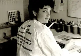 Cathy, with dark curly short hair wearing a paper painters’ suit, sits at a desk and looks over her shoulder at the camera. There is text printed on the back of the suit which reads: BFA, I had the choice (privilege) - most don't, $150 a month for 10 years could buy 50 new bicycles, or a new car, or pay off this education. The image is in black and white. 