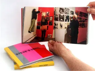 Two hands hold a square artists book, open to images of Cathy installing the Steve’s Vinyl exhibition, putting vinyl albums up onto colourfully painted gallery walls. This open book hovers above two other copies, their cover’s are boldly striped like the gallery walls with yellow, grey, pink and red with Steve’s Vinyl written in all caps in black font in the centre.