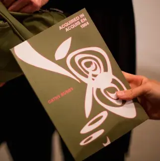 A hand holds a mossy green paperback book with sinuous white shapes rimmed with pink dancing across the cover. Acquired in/Acquis en 1964 is written in the top right corner in matching pink rimmed font. Cathy Busby is written in pink in the middle of the cover.