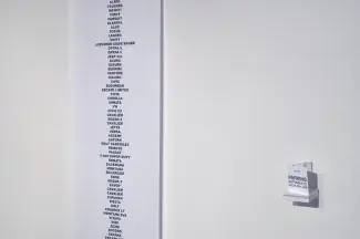 A tiny shelf holds paper folders which read: PARKING GOTTINGEN ST. JULY 8, 2011, 2PM. To the right hanging on the white gallery wall is a long strip of white paper stretched from floor to ceiling listing car names in black sans serif capitals: ALERO, TOUAREG, INFINITI, FORTE, PURSUIT, ELANTRA, SAAB, FOCUS, LANCER, SWIFT, EXPLORER EDDIE BAUER, OPTRA 5, OPTRA 5, JEEP 4X4, ACURA, SATURN, SUNFIRE…which continue out of sight. 