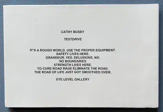 A white rectangular paper back rests on a blue cloth. There is a stack of centre aligned sans serif text running down the middle of the page which reads: CATHY BUSBY, TESTDRIVE. IT’S A ROUGH WORLD. USE THE PROPER EQUIPMENT. SAFETY LIVES HERE. GRANDEUR, YES, DELUSIONS, NO. NO BOUNDARIES. STRENGTH LIVES HERE. TO SURE ROAD RAGE ELIMINATE THE ROAD. THE ROAD OF LIVE JUST GOT SMOOTHED OVER. EYELEVEL GALLERY