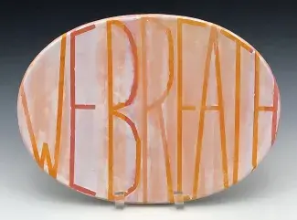 An ovular ceramic platter painted in pink, orange and white rests on a stand. WE BREATH is written in thin tall lettering which bends and stretches, pressing against the borders of the plate at every edge. 