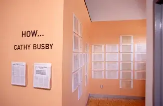 How… Cathy Busby is written on a peach toned gallery wall in the forefront. The peach wall which continues to wrap around into the space on the right is lined with printed pages of scanned self-help books. 