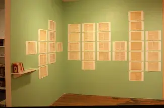 The green walls of a gallery are lined with scans of book pages printed and pinned to the walls in grids. A small white bookshelf installed on the wall holds about 10 self-help books between two book ends. 