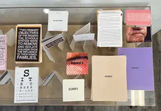 An array of colourful publications by Cathy Busby are laid out on top of a clear vitrine. Some of the books lie flat, including four entitled SORRY in varying sizes and colours, and one which looks like an eye examiners test chart with letters decreasing in size as they descend the page. A few smaller folders stand up partially opened amongst these books. 