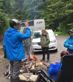 A man in a royal blue rain coat stands with his back to the camera pointing up towards the sky. In front of him there is an open fire pit where a few people are gathered to warm themselves in this clearing in the dense forest. There is a white van and a large passenger bus parked on the dirt road behind them. 