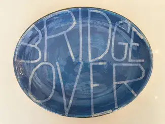 An ovular ceramic platter painted in a wispy grey blue holds the words BRIDGE OVER. These thin tall letters were revealed through tape masking to show the white paint underneath and they bend and stretch to fill the plate.