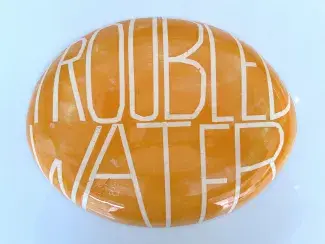 The backside of the ovular ceramic platter is painted in a light creamy orange and white. TROUBLED WATER is written in thin tall tape-maksed letters which reveal the white under paint and swell to fit the plates' convex shape of the plate.