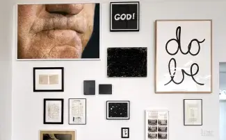A brightly lit white gallery wall is filled with art works. A large print of a mouth biting its lips overlooks the works from the top left corner. Below it are a series of framed archival documents and two small canvases covered in woven film rolls. To the right of the mouth a black print reads GOD! in white text, below it is a canvas with black rope mark scribbles and a framed print that looks like a constellation. A large framed white print reads DO BE in black looping cursive on the far right. 