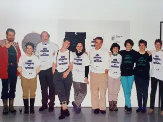 Ten people of varying heights and genders stand in front of a gallery installation. Each one is wearing an oversized white or black t-shirt on top of their other clothes which reads ANNA LEONOWENS WAS A SUFFRAGIST in purple or green capital lettering. Cathy Busby stands in the middle of the line of folks smiling at the exhibition she and her staff put together.