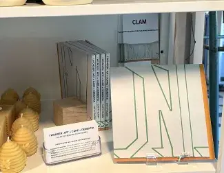 A square book is on display on a cozy cafe shelf alongside beeswax candles. The cover of the book is white with abstract letter-looking outlines in green painters tape. 