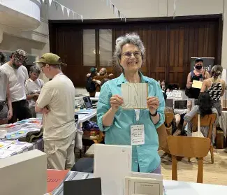 Cathy smiles widely while holding a copy of I WONDER: Art + Care + Dementia up to her chest. She is standing at her table at the Halifax Art Book Fair and people are milling around the vendors throughout the room. 