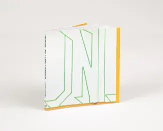 A small square book rests upright at an angle, the spine reads: I WONDER: ART + CARE + DEMENTIA CATHY BUSBY AM. The cover of the book is white with abstract letter-looking outlines in green painters tape. 