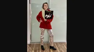 Cathy stands proudly in metallic silver pants and her fathers red knit Cowichan style sweater. She is holding sheet music for The Kinks song Lola in one hand while the other hand rests on her hip. She wears silver earrings and red lipstick which match her outfit. 