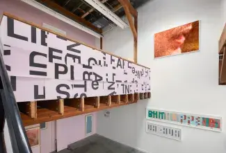 A large framed print of an open mouth of a politician looms over the gallery space. Another mouth print, smaller in size, hangs on the lower wall adjacent to four Atrium prints, silhouettes of vitrines and artworks in blue and gold. WE ARE SORRY Fragments pattern the mezzanine wall, their yellowing-pink colour toning with the pink and purple wall paint which divides the gallery space.