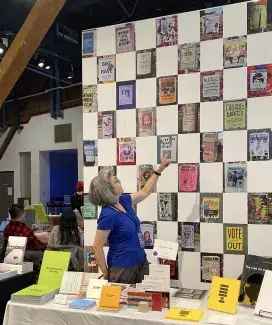  Cathy Busby, wearing a blue t-shirt and her grey hair down around her shoulders, points up to her wall of poster photos which frame her colourful table of publications at the Vancouver Art Book Fair. 