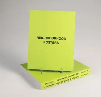 A large bright green book entitled NEIGHBOURHOOD POSTERS in bold black lettering rests on a book stand atop two other copies of the book. The book spines read: CATHY BUSBY NEIGHBOURHOOD POSTERS VANCOUVER.