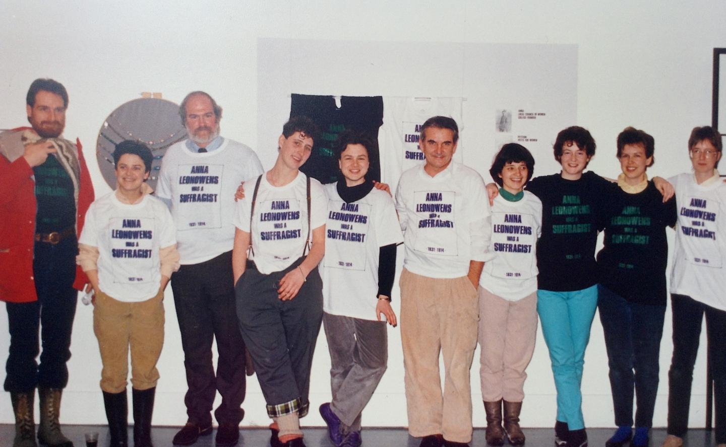 Ten people of varying heights and genders stand in front of a gallery installation. Each one is wearing an oversized white or black t-shirt on top of their other clothes which reads ANNA LEONOWENS WAS A SUFFRAGIST in purple or green capital lettering. Cathy Busby stands in the middle of the line of folks smiling at the exhibition she and her staff put together.