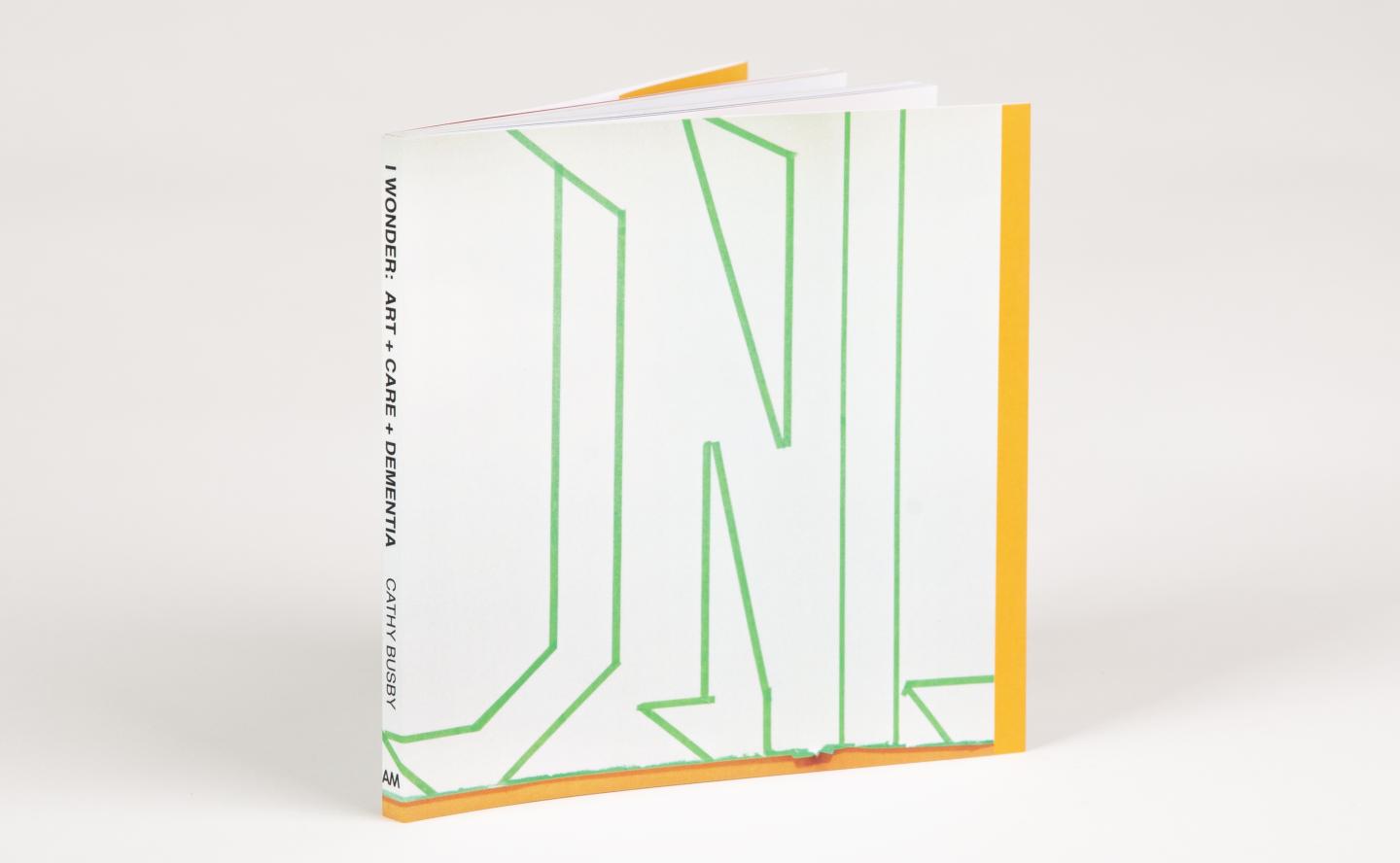 A small square book rests upright at an angle, the spine reads: I WONDER: ART + CARE + DEMENTIA CATHY BUSBY AM. The cover of the book is white with abstract letter-looking outlines in green painters tape. 